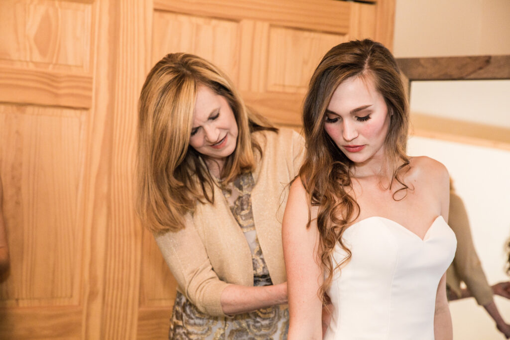 A woman helps another woman adjust her strapless white dress in a warmly lit room with wooden doors.