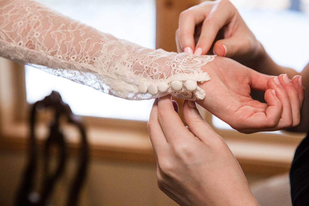 Close-up of hands fastening buttons on a white lace sleeve of a garment, possibly a wedding dress, in a softly lit room.