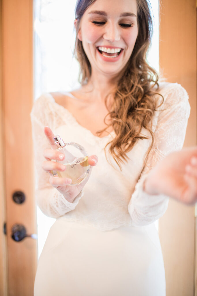 A woman in a wedding dress sprays perfume on her wrist, smiling. She stands in front of a wooden door, holding a round perfume bottle.