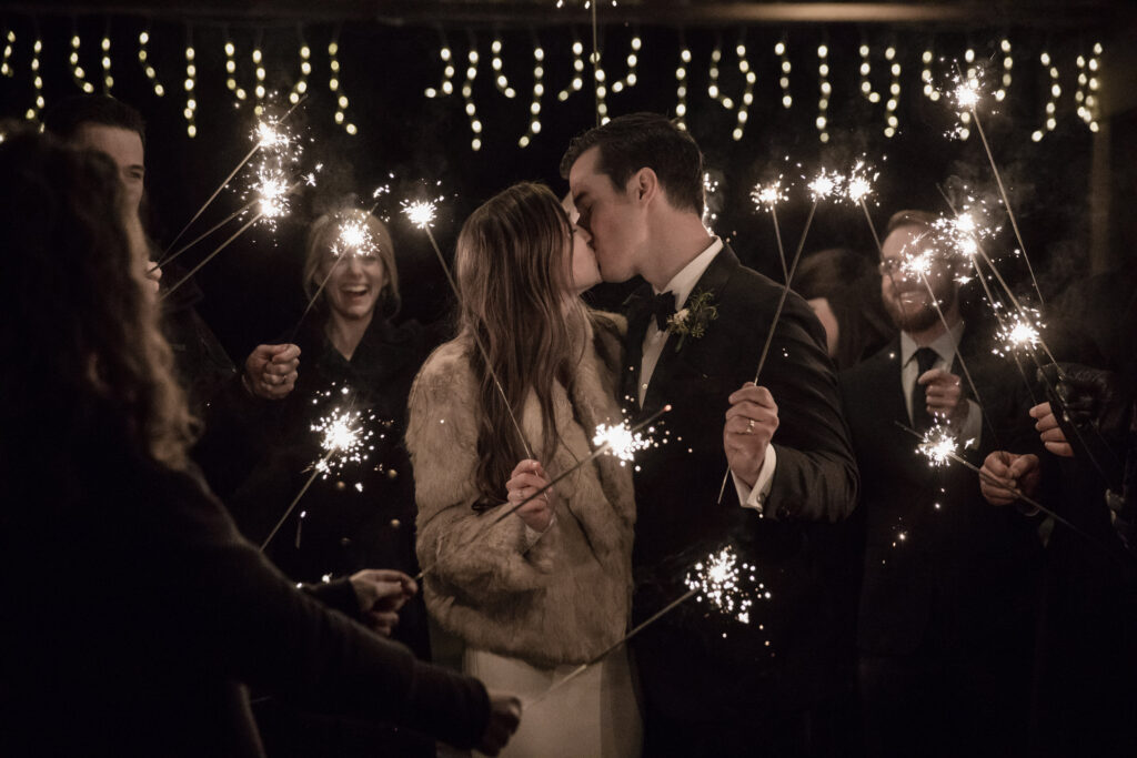 Couple kissing surrounded by people holding sparklers at night under string lights.