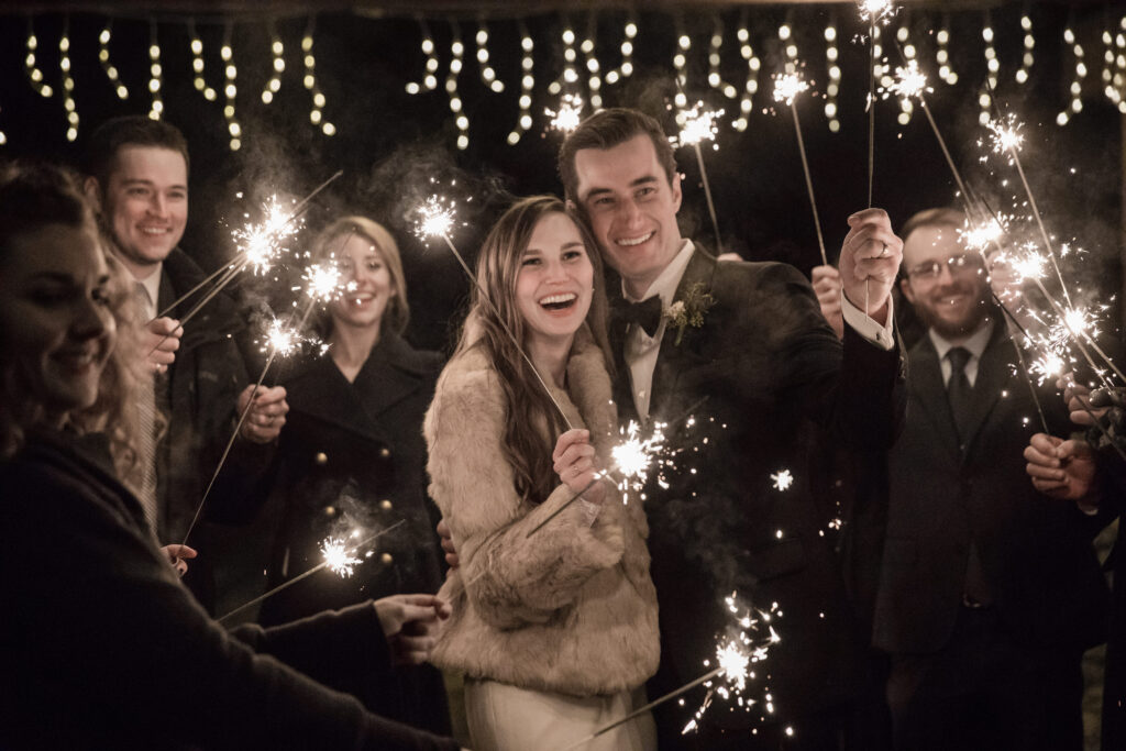 A newlywed couple smiles while surrounded by friends holding sparklers at night. Twinkling lights hang overhead.