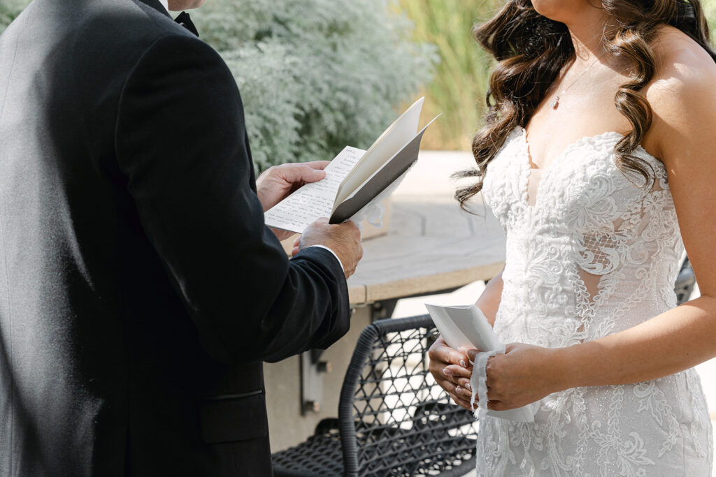 A bride and groom in formal attire exchange vows outdoors, holding papers.