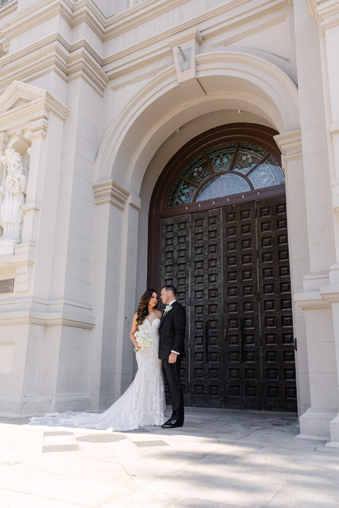 A couple dressed in a wedding gown and suit stand close together in front of a large wooden door on a sunlit stone building.