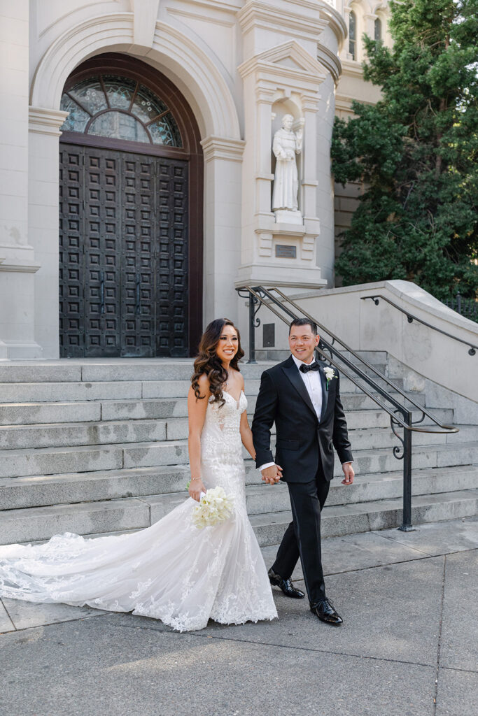 A couple in formal attire holds hands while walking down steps in front of a large building with ornate doors and a statue.
