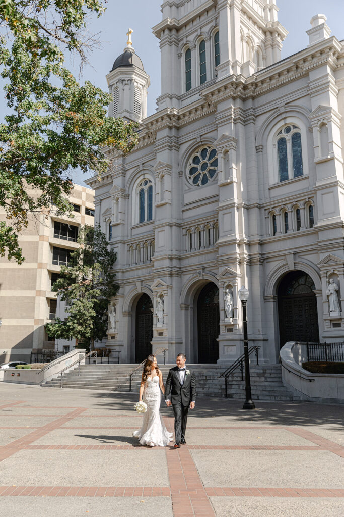 A bride and groom walk hand in hand outside a large, ornate church with arched doors and statues.