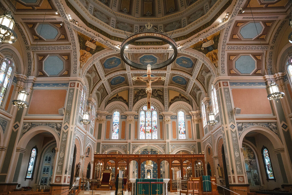 Ornate church interior with decorative arches, a large cross hanging from the ceiling, stained glass windows, and chandeliers illuminating the space.
