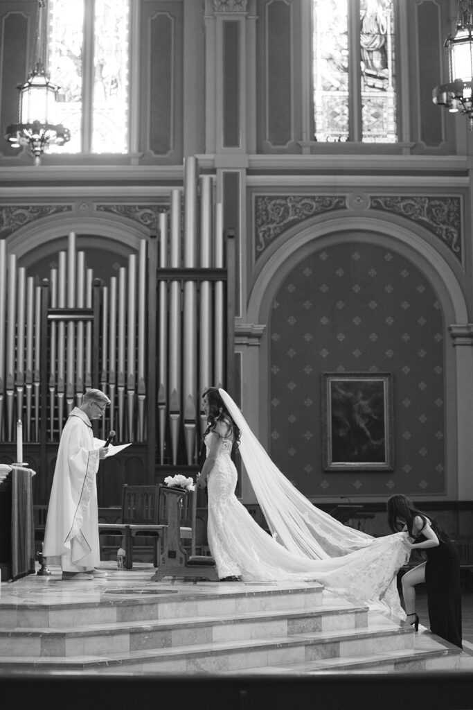 Bride and groom stand at the altar during a wedding ceremony in a church. A woman adjusts the bride's long train.