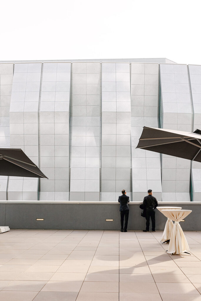 Two people stand on a terrace, looking at a modern building facade with a geometric pattern. Umbrellas and covered tables are visible in the foreground.