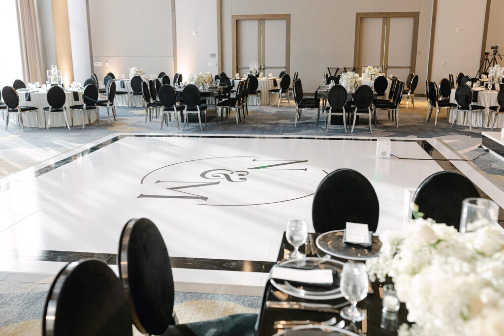 Spacious dining area set for an event with round tables, white tablecloths, black chairs, and a decorative dance floor with initials.