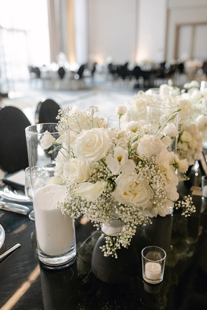 Elegant table centerpiece with white roses, baby's breath, and tall glasses on a black table. Candles and chairs are visible in the softly lit background.