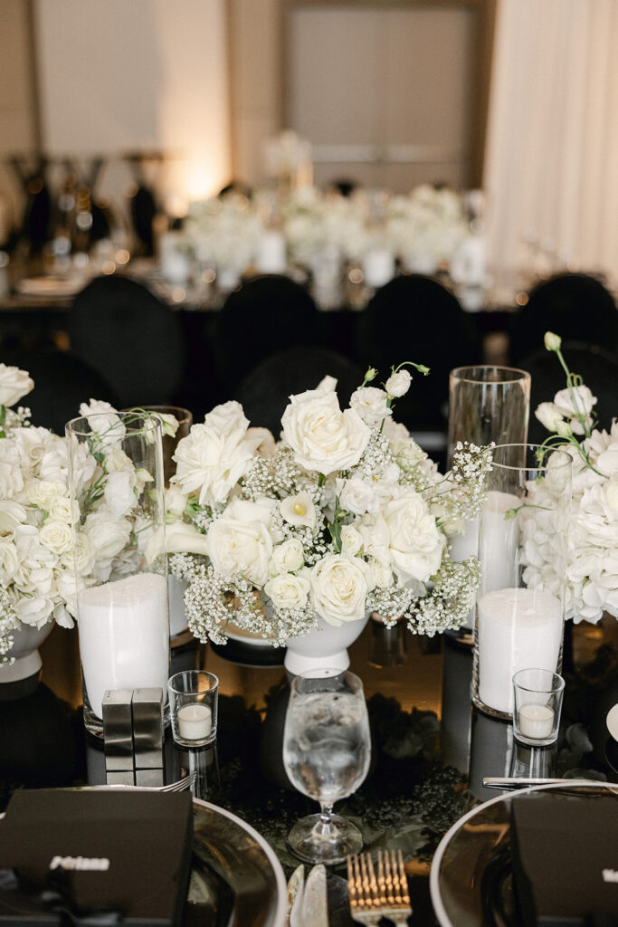 Elegant table setting with white rose centerpieces, surrounded by candles and glassware on a dark reflective surface.