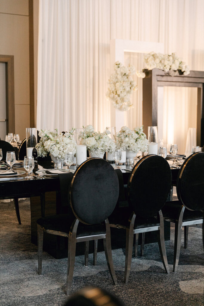 Elegant dining table setup with black chairs, white floral centerpieces, and place settings in a room with white drapes and floral decor.