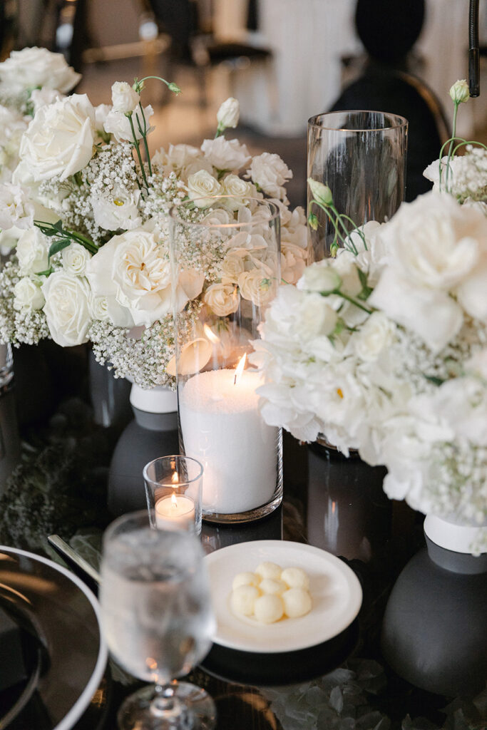Elegant table setting with white floral arrangements, a lit candle in a glass holder, a glass of water, and a plate with small white spheres on a glossy black surface.