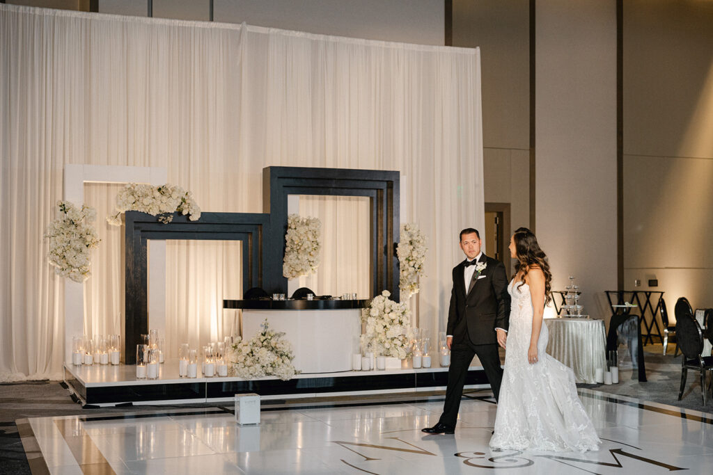 A bride and groom walk on a white glossy floor in front of a modern altar with black frames, white flowers, and candles, perfectly orchestrated by a Sacramento wedding planner.