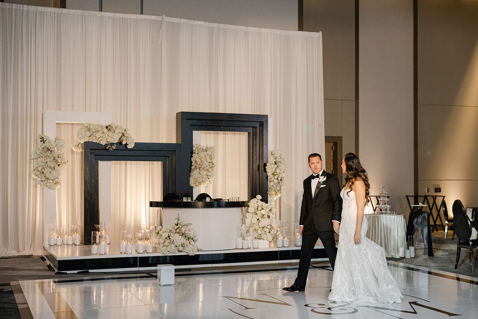 A bride and groom walk on a white glossy floor in front of a modern altar with black frames, white flowers, and candles, perfectly orchestrated by a Sacramento wedding planner.
