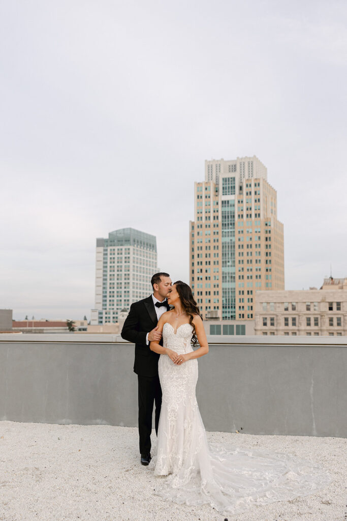 A couple in formal attire stands on a rooftop with city buildings in the background. The woman wears a white gown, and the man is in a black suit.