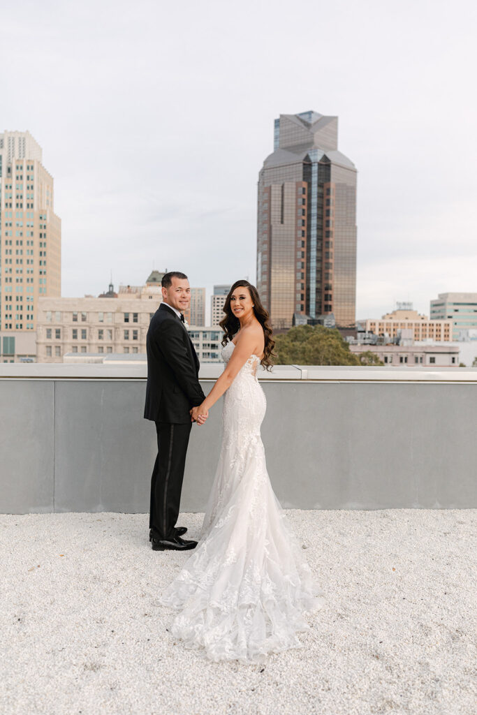 A couple in formal wedding attire stands on a rooftop holding hands, with city skyscrapers in the background.