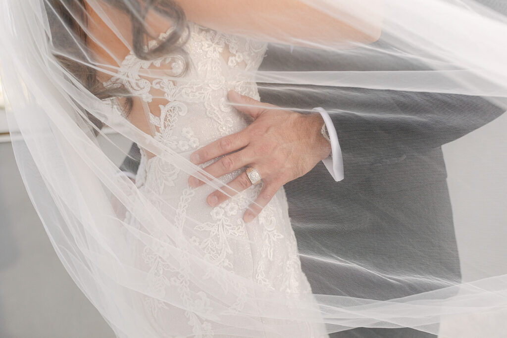 A bride and groom embrace under a sheer veil, captured perfectly by their Sacramento Wedding Planner. The bride's lace dress and the groom's suit are partially visible, with his hand resting on her back, highlighting a glimmering wedding ring.