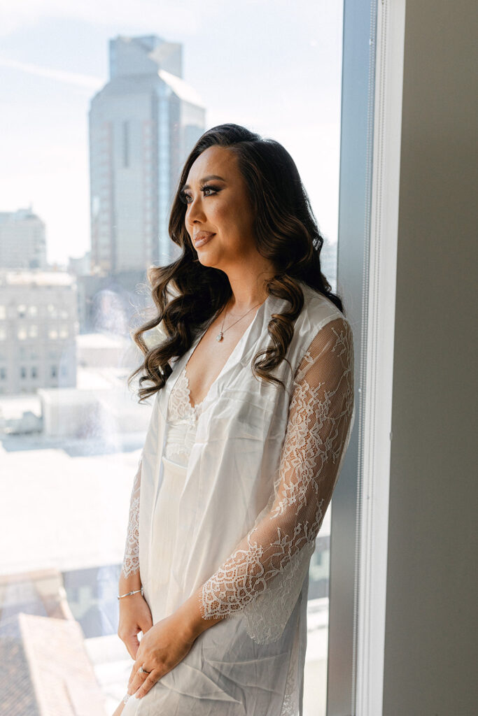 A person with long hair in a white lace robe stands by a window, overlooking a cityscape with buildings in the background.