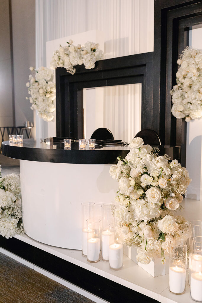 Elegant white and black DJ booth with surrounding white floral arrangements and lit candles in glass holders, set against a backdrop of draped white fabric.