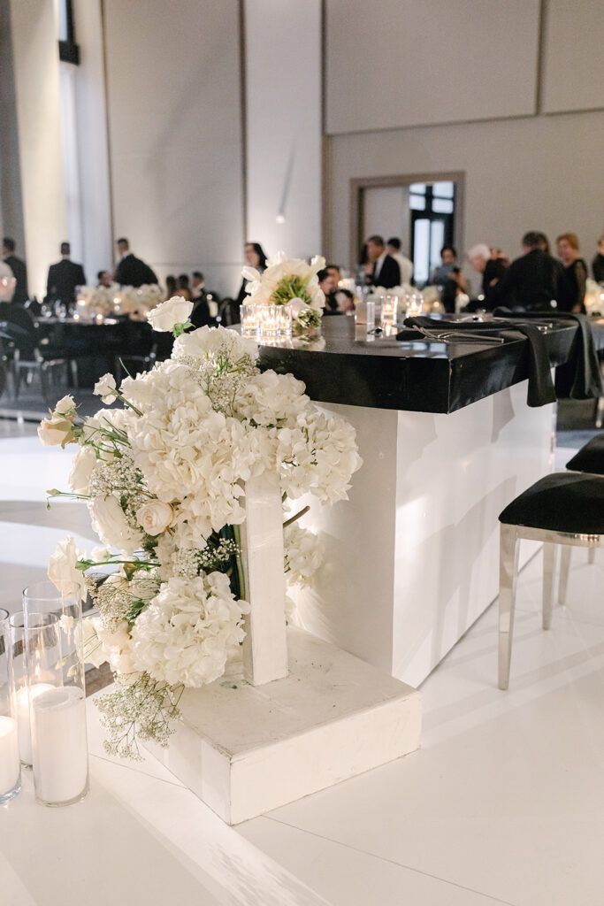 Elegant event setting with white floral arrangements on a pedestal, black and white decor, candles, and guests seated at tables in the background.