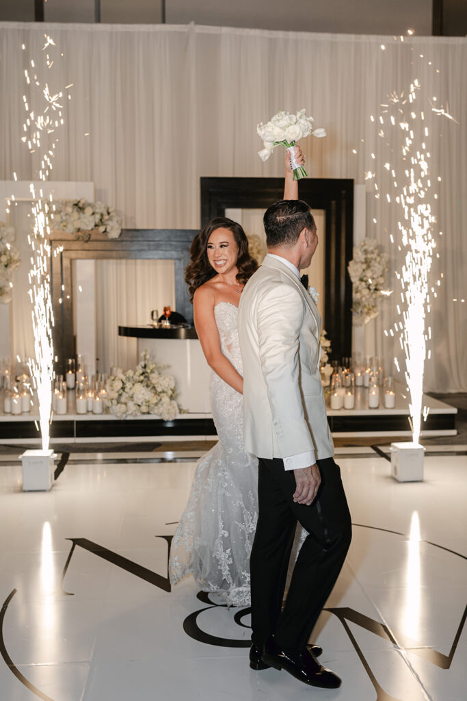 A bride and groom dance on a white floor with initials, surrounded by fireworks and floral decorations. The bride holds a bouquet aloft, smiling.