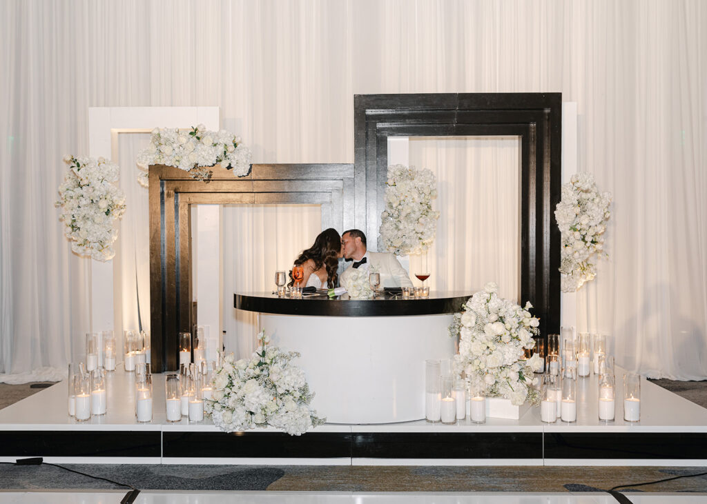 A couple sits at an elegantly decorated table with large black and white frames and white floral arrangements.