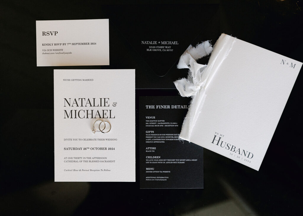 A wedding invitation set with RSVP card, envelope, details card, and a small book labeled "To My Husband." Cards display names Natalie & Michael and event information. Rings are placed on the main card.