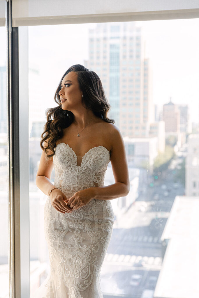 Bride in a strapless lace wedding gown stands by a window, gazing outside. City buildings are visible in the background.