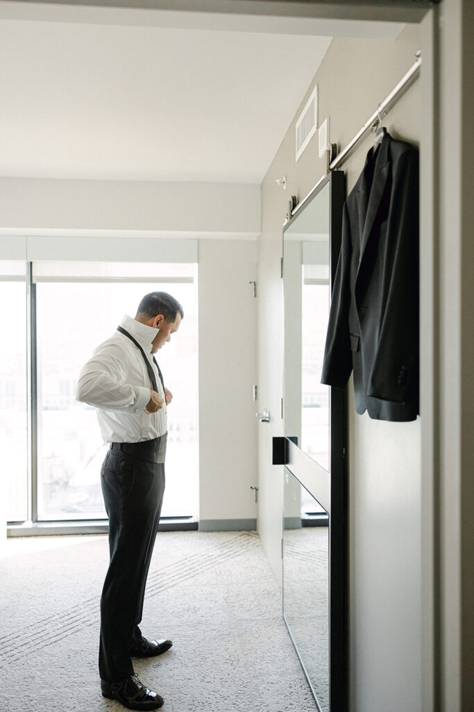 Man in a hotel room adjusting his suspenders, with a suit jacket hanging nearby on a door handle.
