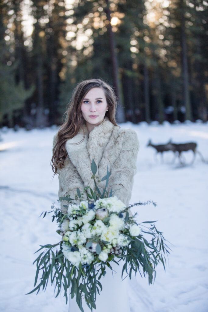 A woman in a fur shawl holds a bouquet of white flowers and greenery in a snowy forest with deer in the background.