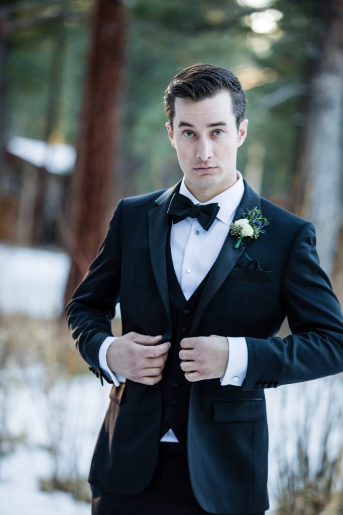 Man in a black tuxedo with a bow tie and boutonniere stands in a snowy, forested area, adjusting his jacket.