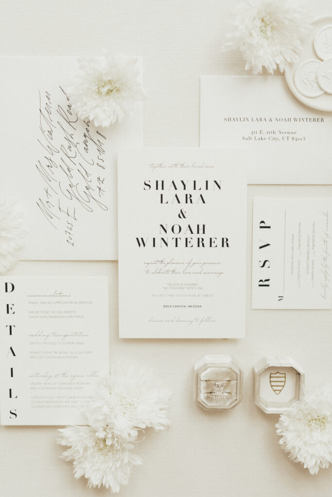 Flat lay of wedding invitations and RSVP cards arranged with white flowers, featuring the names Shaylin Lara and Noah Winterer, with rings in boxes near the bottom.