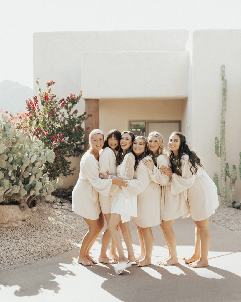 Six women wearing matching light robes stand together, embracing and smiling, outside a building with desert plants around them.