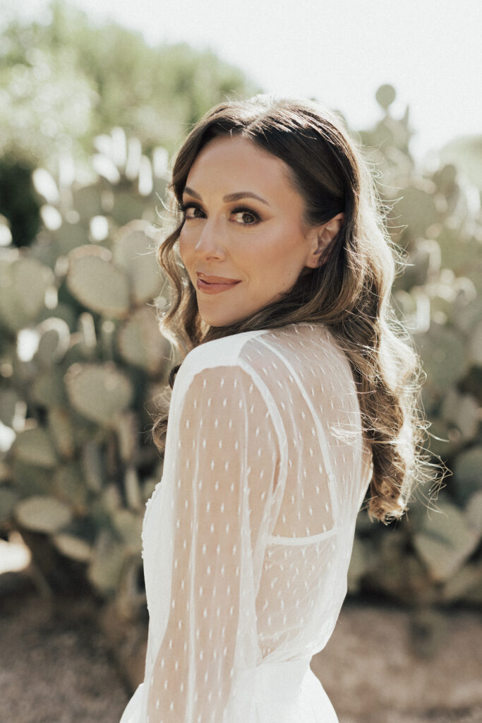 A woman in a white, semi-sheer dress with long, wavy hair stands in front of a cactus plant, looking over her shoulder.