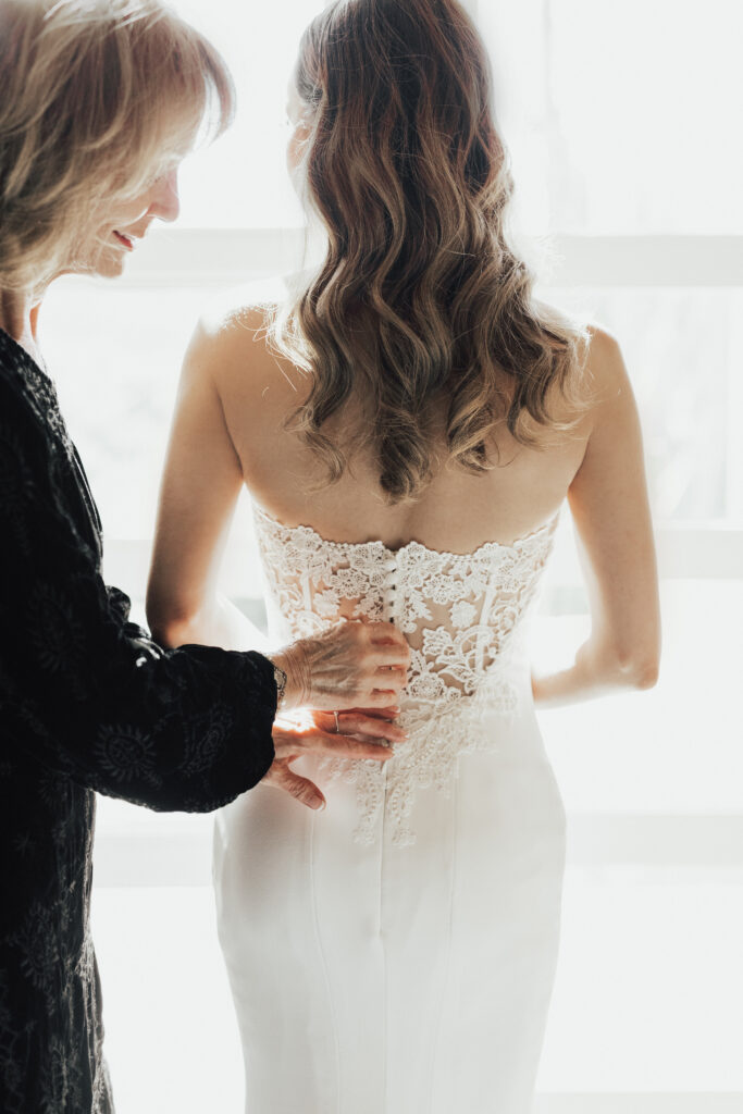 A woman helps another woman button up the back of her lace wedding dress in front of a bright window.
