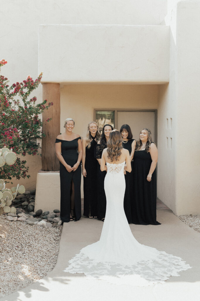 A bride in a white dress stands facing five bridesmaids in black dresses outside a building with desert landscaping.