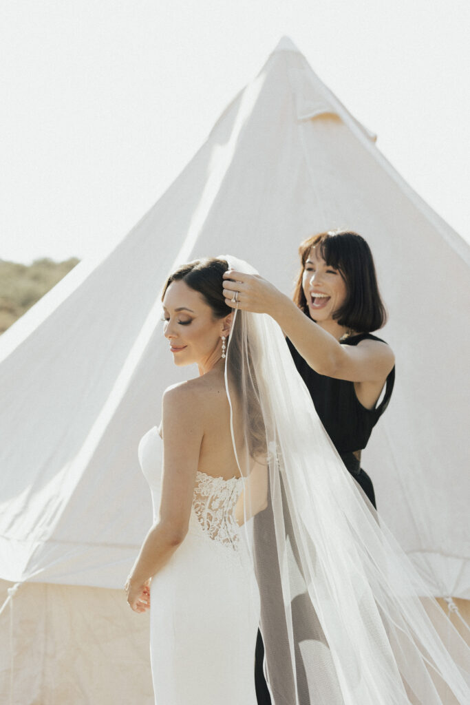 A person in a white wedding dress stands with eyes closed, while another person adjusts the veil against a tent backdrop.