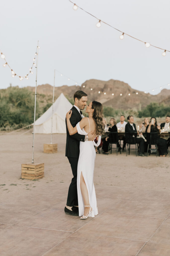 A couple dances on an outdoor wooden floor with string lights overhead. A small group of people is seated at a table in the background, with mountains visible in the distance.