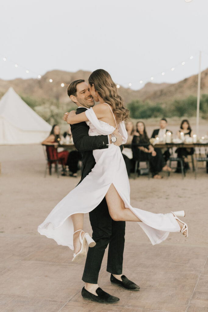 A man in a suit lifts and spins a woman in a white dress outdoors, with a group of seated people and string lights in the background.