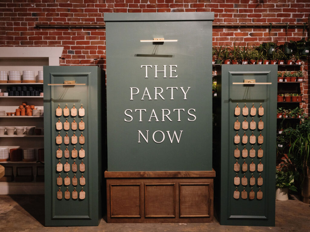 A green display reads "The Party Starts Now" against a brick wall backdrop, flanked by panels with hanging tags. Shelves adorned with pottery and plants add charm to this luxurious setting, curated by a top Sacramento event planner.