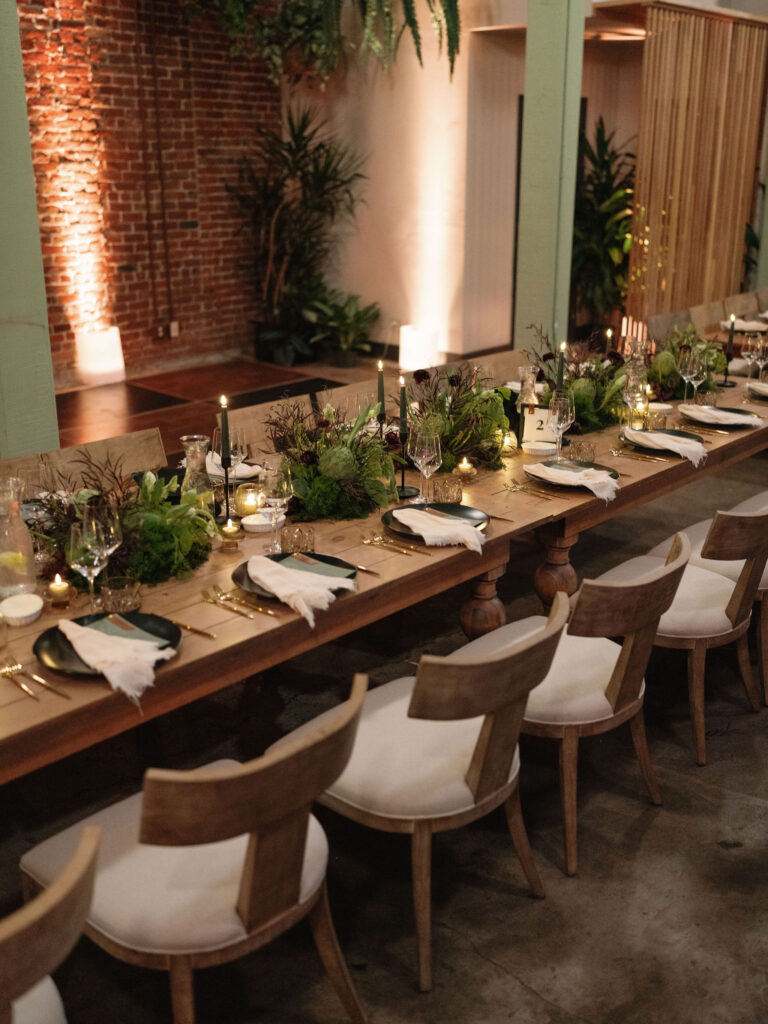 A long wooden table set for dinner with white chairs, green and white floral centerpieces, candles, and folded napkins. Brick wall and plants in the background.