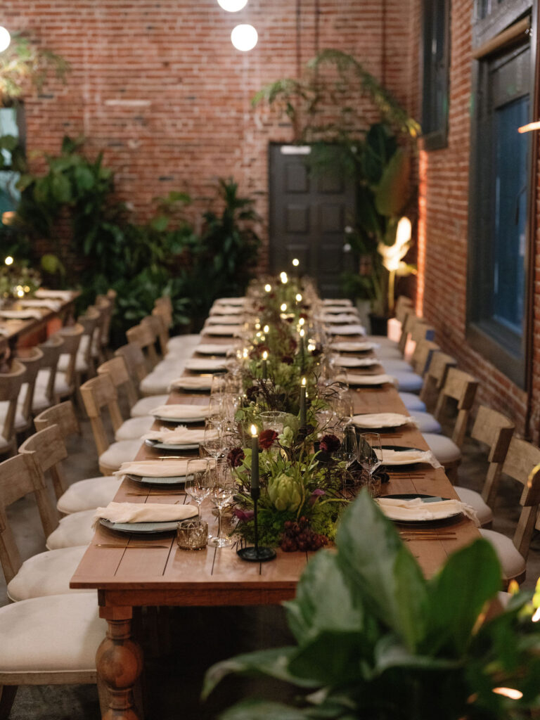 Elegant dining setting with a long wooden table, white plates, wine glasses, and floral centerpiece. Surrounded by wooden chairs, brick walls, and green plants under soft lighting.
