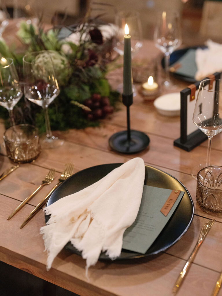 Elegant table setting with a black plate, white cloth napkin, menu, gold cutlery, wine glasses, candle in a black holder, and a centerpiece of greenery and grapes on a wooden table.