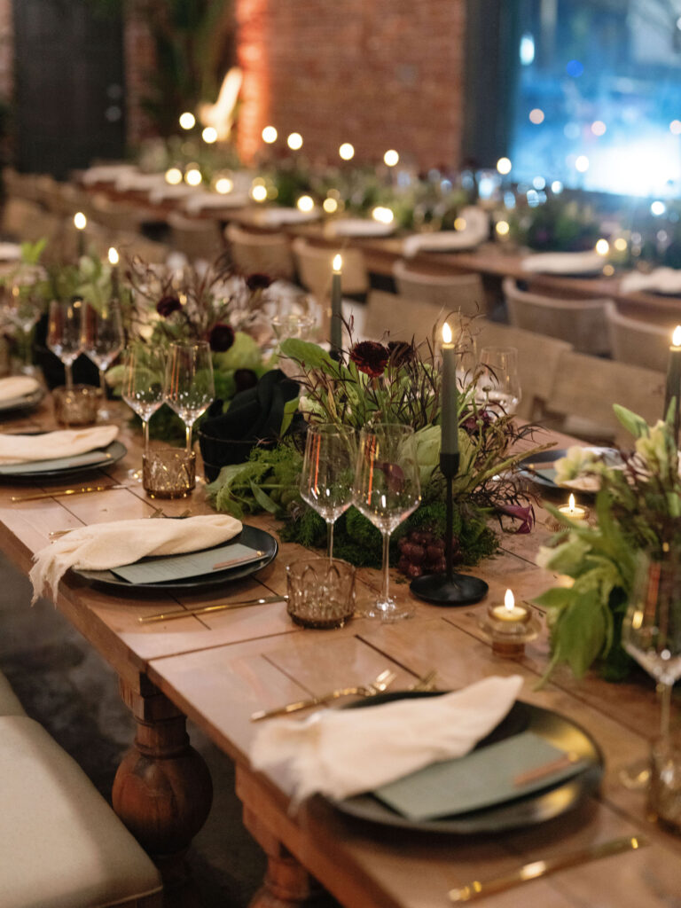 Elegant dining table set for an event with candles and greenery, featuring neatly arranged plates, napkins, and glasses.