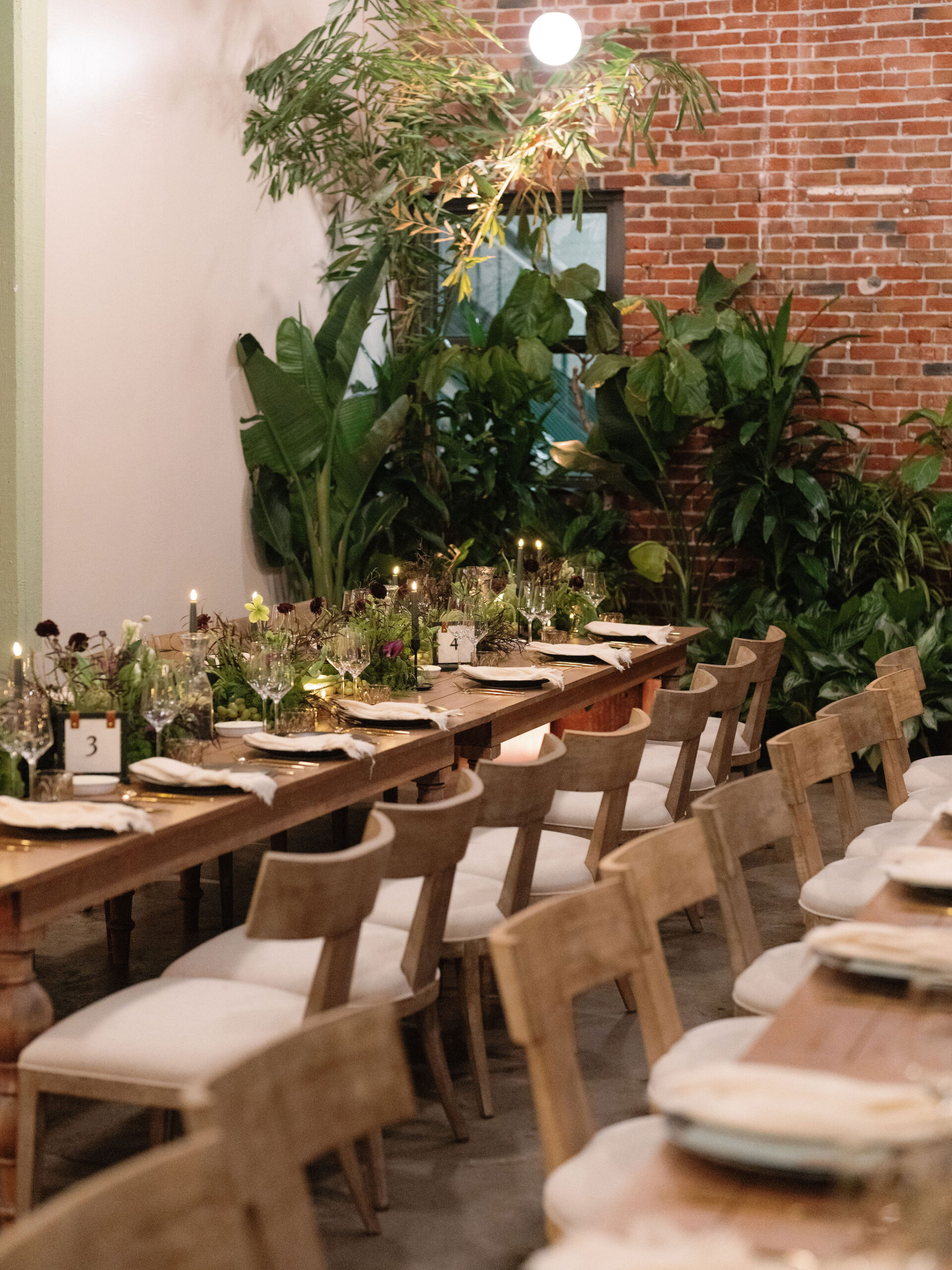 Long wooden dining tables with white cushioned chairs are set with glassware and greenery. The setting includes brick walls, plants, and soft lighting.