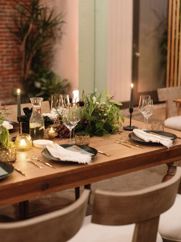 A wooden dining table set for a meal with wine glasses, plates, cloth napkins, candles, and a greenery centerpiece.