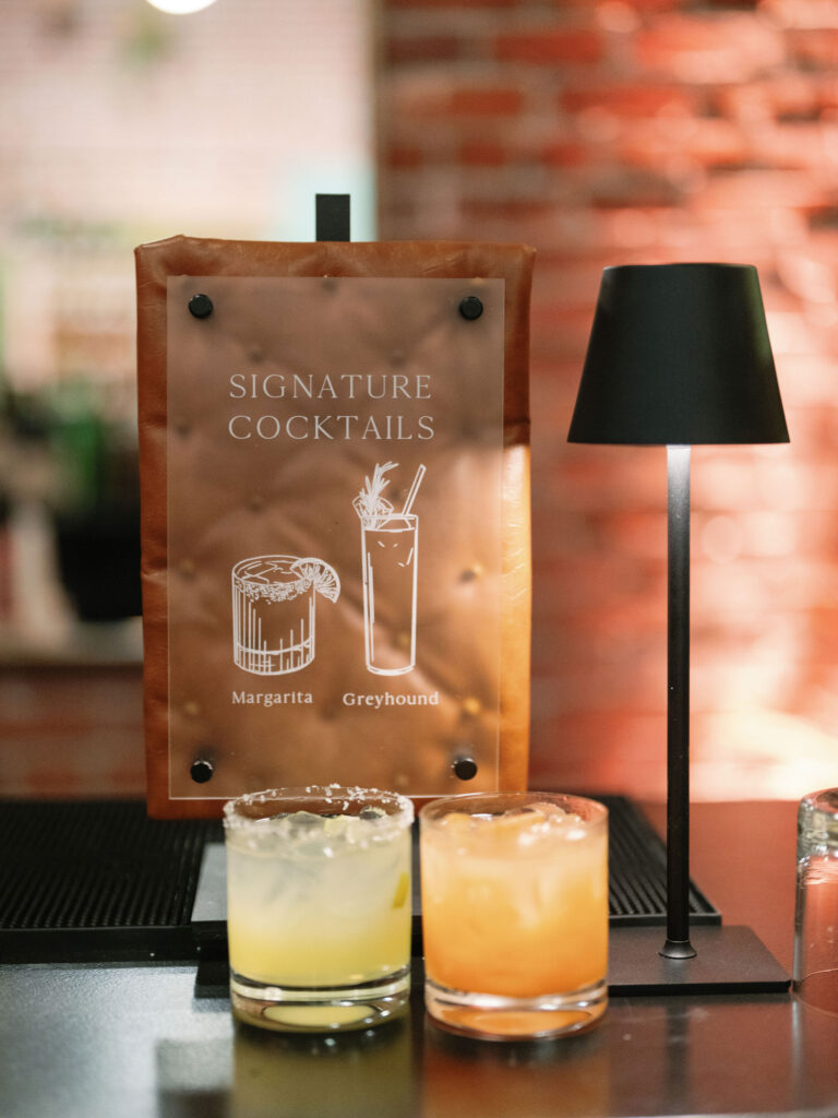 Sign listing two cocktails, Margarita and Greyhound, next to a small black lamp. Two glasses with yellow and orange drinks in the foreground. Brick wall in the background.