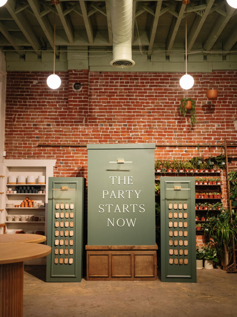 Green decorative setup with the words "The Party Starts Now" on a brick wall. Flanked by displays with plants and small hanging items. Two hanging lights above.
