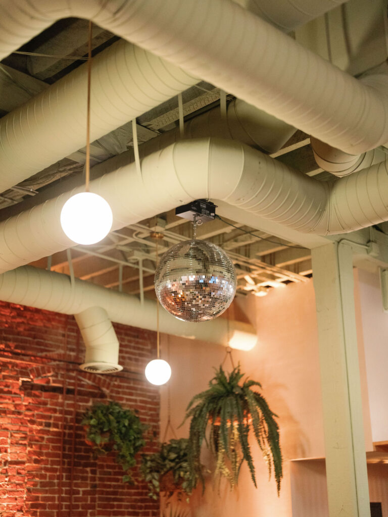 A disco ball hangs from the ceiling alongside round lights, with exposed ducts and a brick wall adorned with hanging green plants in the background.